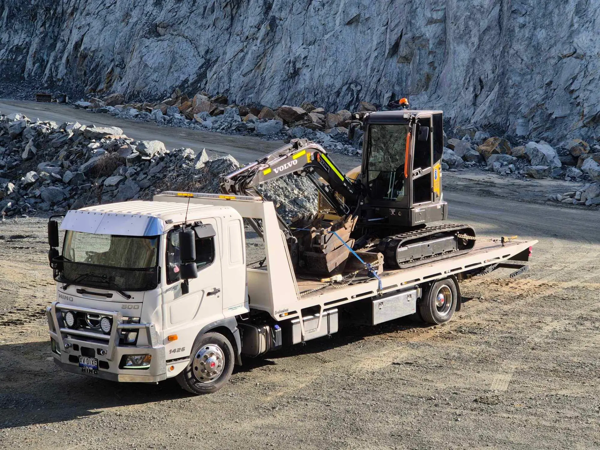 White truck carrying an excavator with a quarry in the background